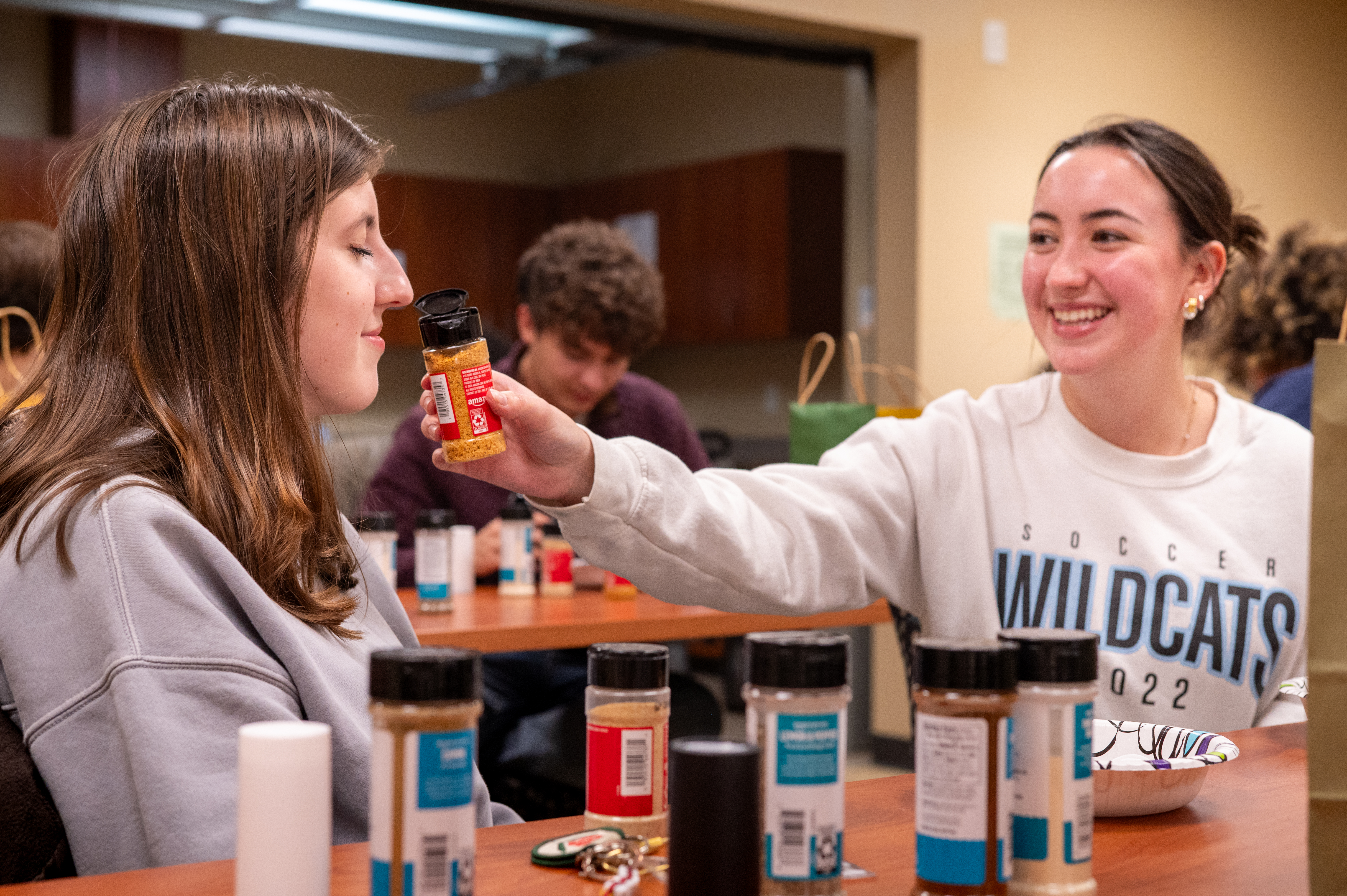 Two students sit at a classroom table during a lab activity, with one student holding a small container near the other’s face as if conducting a scent or observation test. Several labeled jars and supplies are arranged on the table between them.