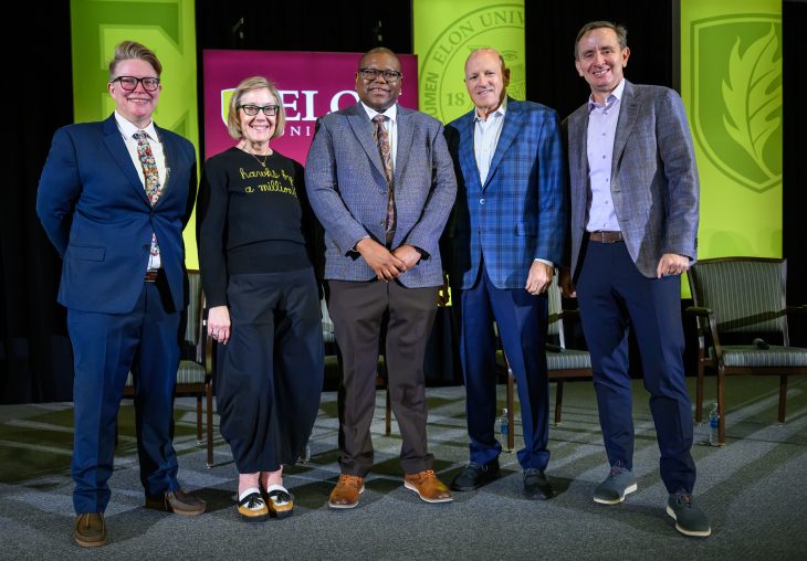 Five panelists stand on stage at Elon University’s Winter Symposium, smiling for a group photo in front of green Elon banners.