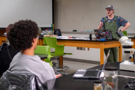 A student presents at the front of a classroom lab while classmates sit at tables with laptops and microscopes. The presenter gestures while speaking, and green chairs and lab equipment are visible throughout the room.