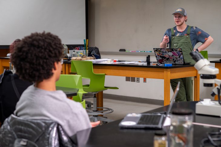 A student presents at the front of a classroom lab while classmates sit at tables with laptops and microscopes. The presenter gestures while speaking, and green chairs and lab equipment are visible throughout the room.