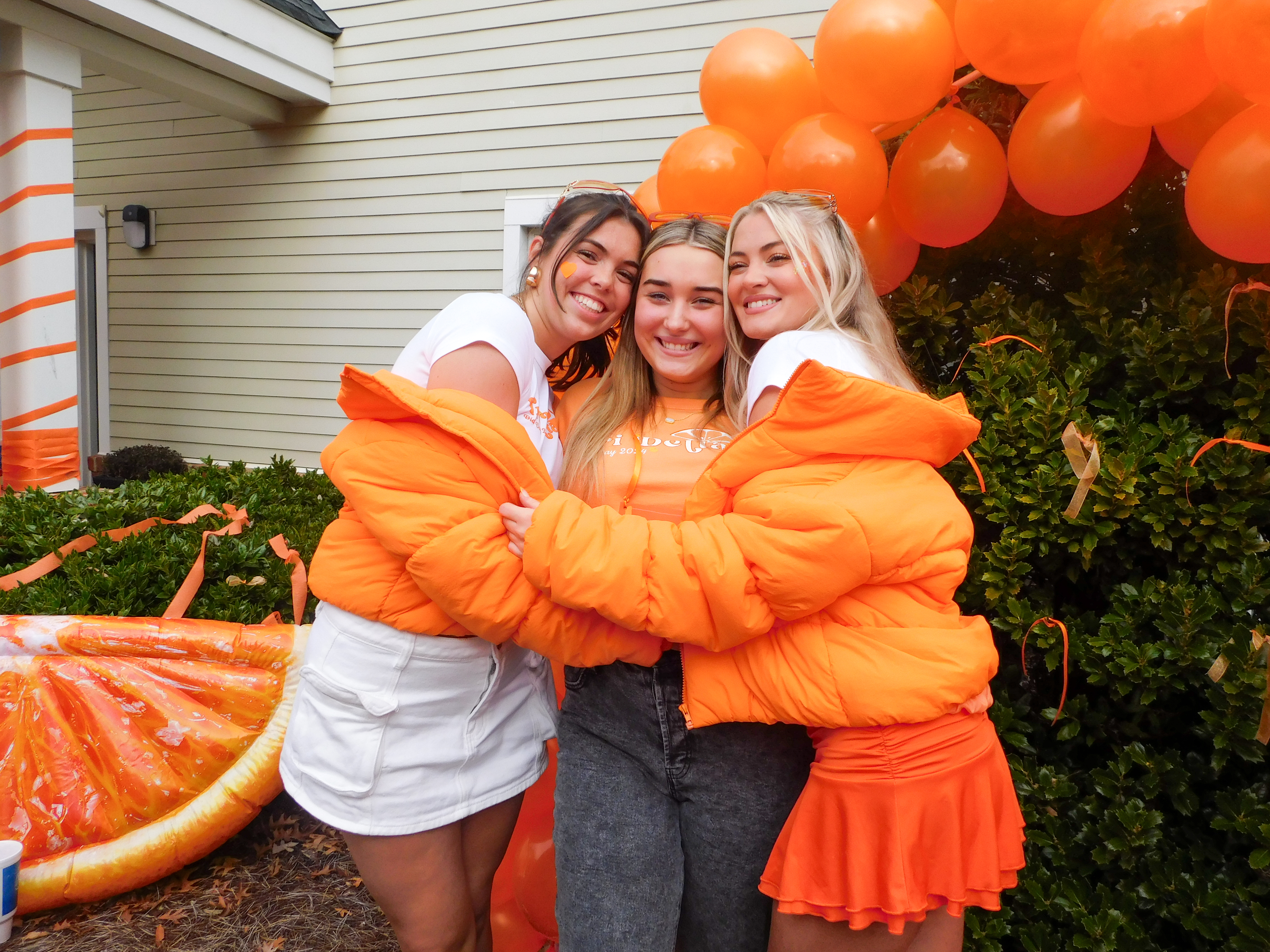 Three individuals in matching orange puffer jackets smiling and embracing at an outdoor event with orange balloons in the background.