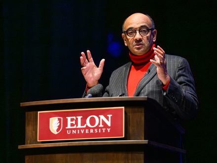 A man with glasses, red turtleneck and grey blazer speaks in front of an Elon University podium