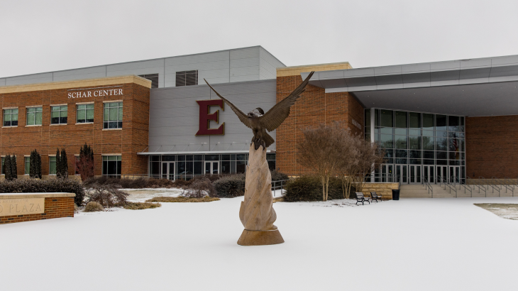 A campus building with brick and modern glass architecture stands behind a sculpture of an eagle with wings spread. Snow covers the ground, and the building signage and large red letter “E” are visible, indicating a university campus setting.