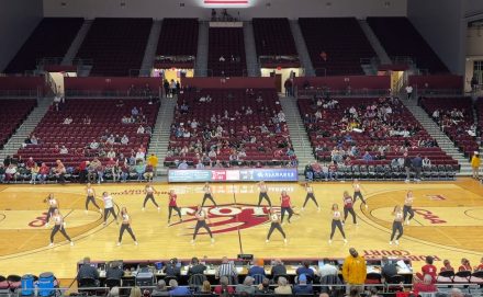 A dance team performs a synchronized routine at center court during a basketball game in a large arena. Spectators sit in the stands surrounding the court, watching the performance.