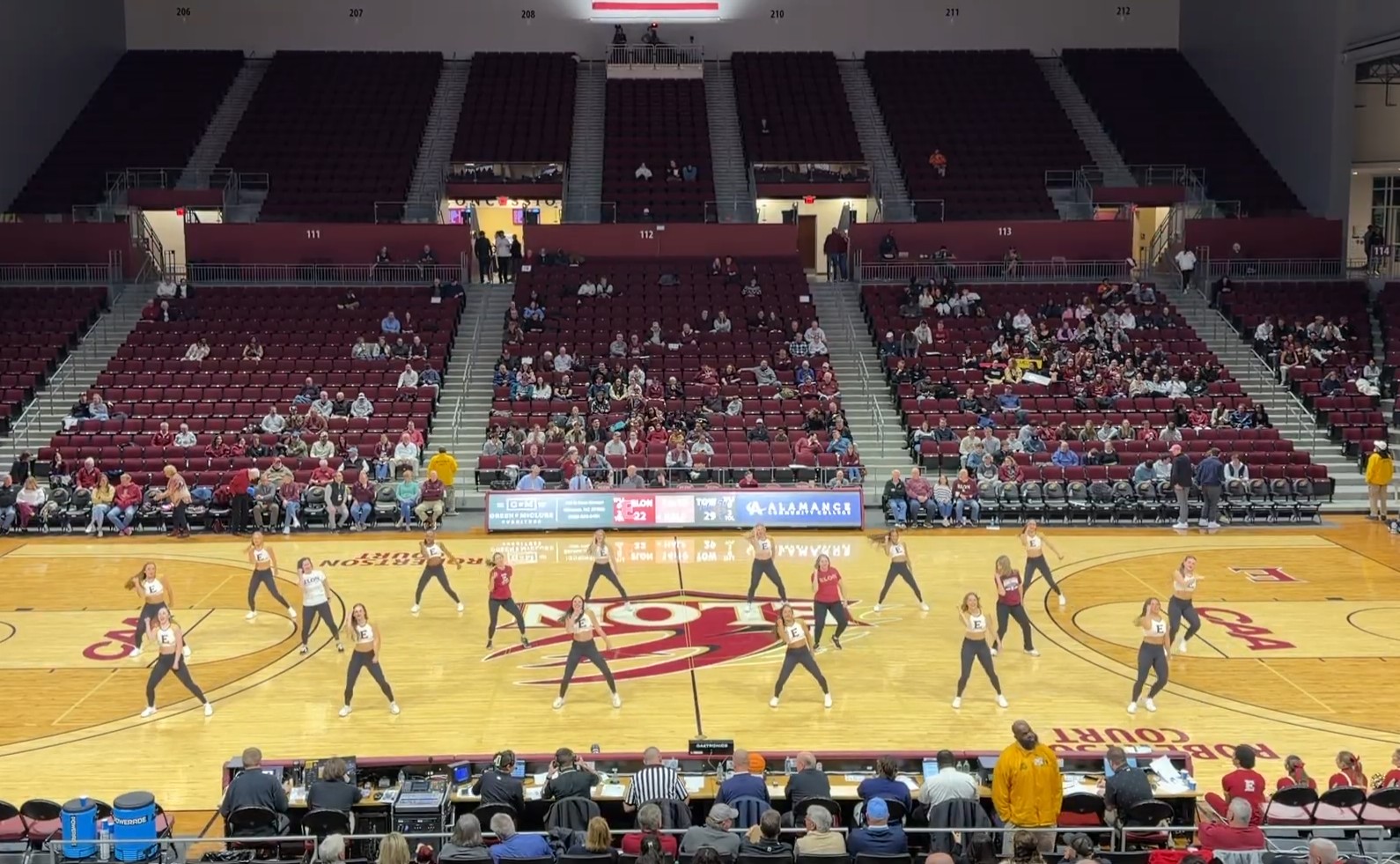 A dance team performs a synchronized routine at center court during a basketball game in a large arena. Spectators sit in the stands surrounding the court, watching the performance.