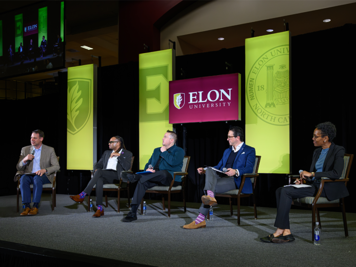 Five panelists sit on stage during Elon University’s Winter Symposium, engaging in discussion beneath green Elon banners and an Elon University screen.