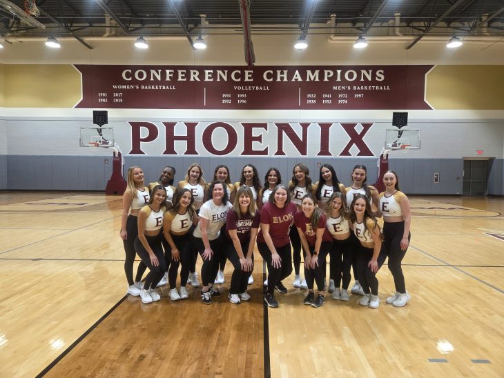 A group of students pose together on a basketball court inside a gymnasium, smiling for a team photo. A large “PHOENIX” sign and championship banners hang on the wall behind them, indicating a collegiate athletic setting.