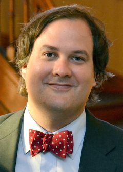 A headshot of Enrique Armijo, smiling, wearing a red bowtie and gray blazer
