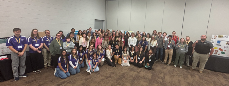A very large group of students, faculty, and staff pose together in a conference room in front of educational poster displays. The group smiles at the camera, wearing name badges, suggesting a formal academic showcase or professional event.