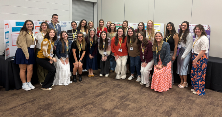A large group of students and presenters pose together in front of multiple educational poster displays at a conference or showcase event. Everyone is smiling and wearing conference badges, indicating a collaborative academic or professional learning setting.