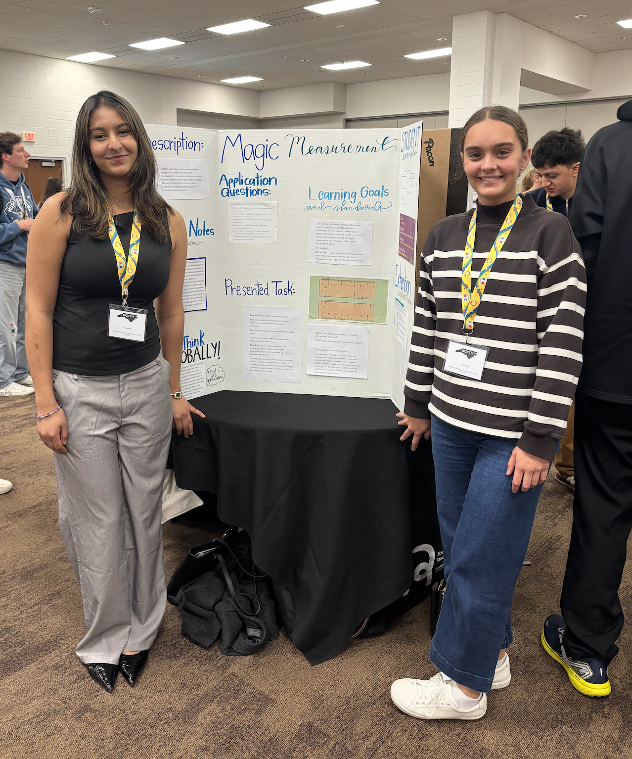Two students stand smiling beside a tri-fold poster titled “Magic Measurement” at an academic poster session. The display includes sections on learning goals, application questions, and a presented task, with other attendees visible in the background.