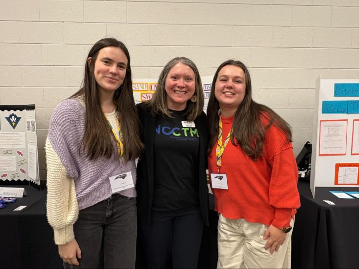 Three presenters stand together smiling at an educational poster session, wearing conference badges. Display boards with instructional materials are visible behind them, indicating a professional learning or academic conference setting.