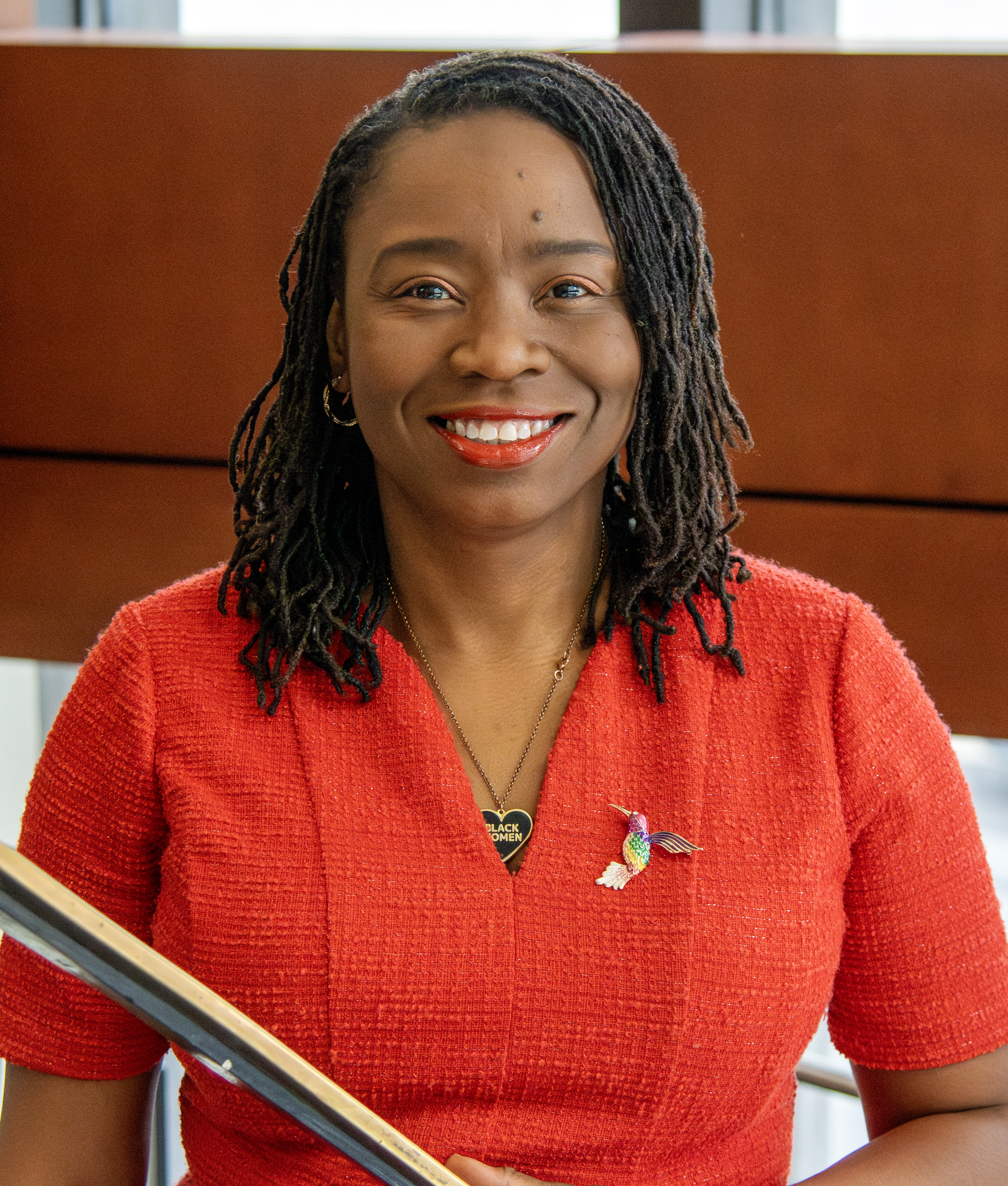 A woman smiles warmly on a spiral staircase at Elon Law. She is wearing a crimson top with a humminbird charm.