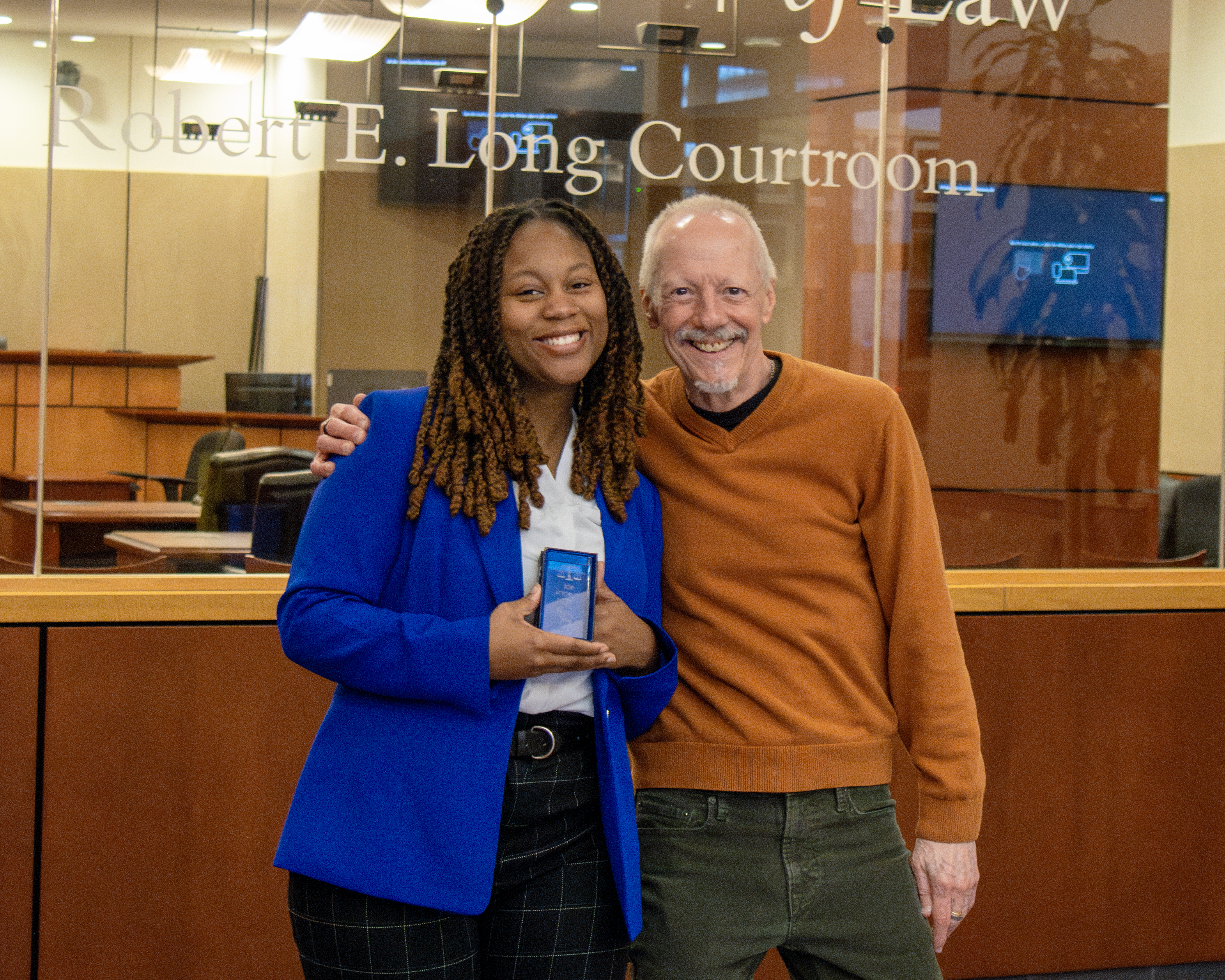 A female law student and male law professor outside the Robert E Long Courtroom at Elon Law. They are smiling. The student is holiding a trophy plaque.