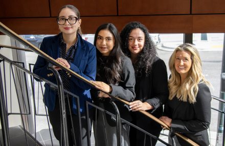Four women in professional dress standing on a spiral staircase. They are smiling at the camera.