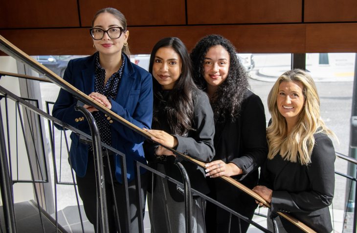 Four women in professional dress standing on a spiral staircase. They are smiling at the camera.