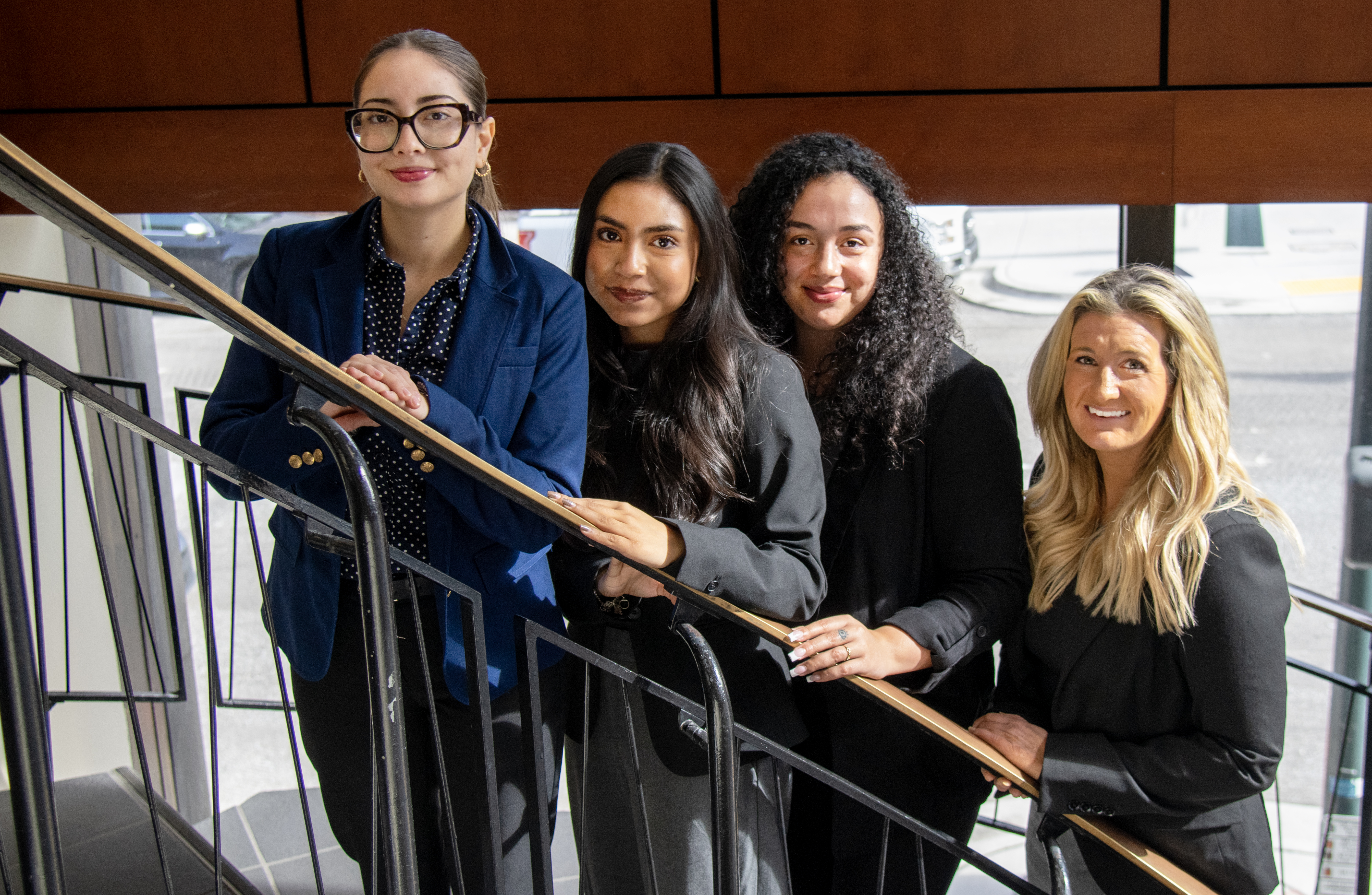 Four women in professional dress standing on a spiral staircase. They are smiling at the camera.