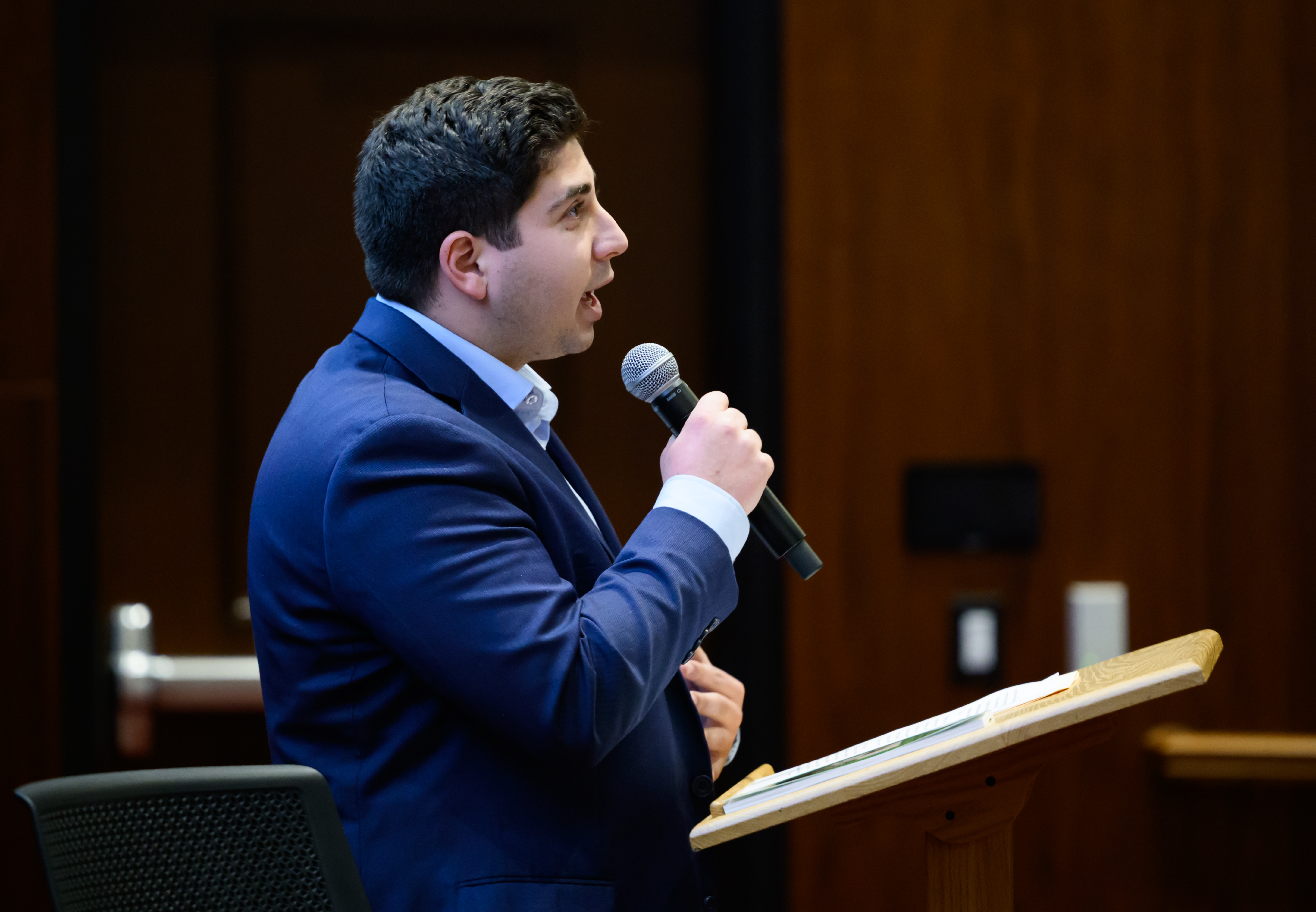 A man in a suit speaks into a microphone at a podium in a lecture hall, addressing an audience. 