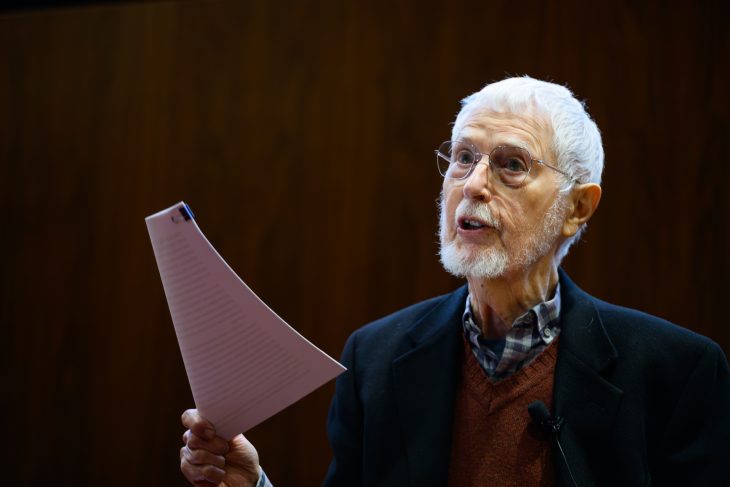 An older man with white hair and a beard speaks while holding a sheet of paper, seated at the front of a room with a microphone nearby.