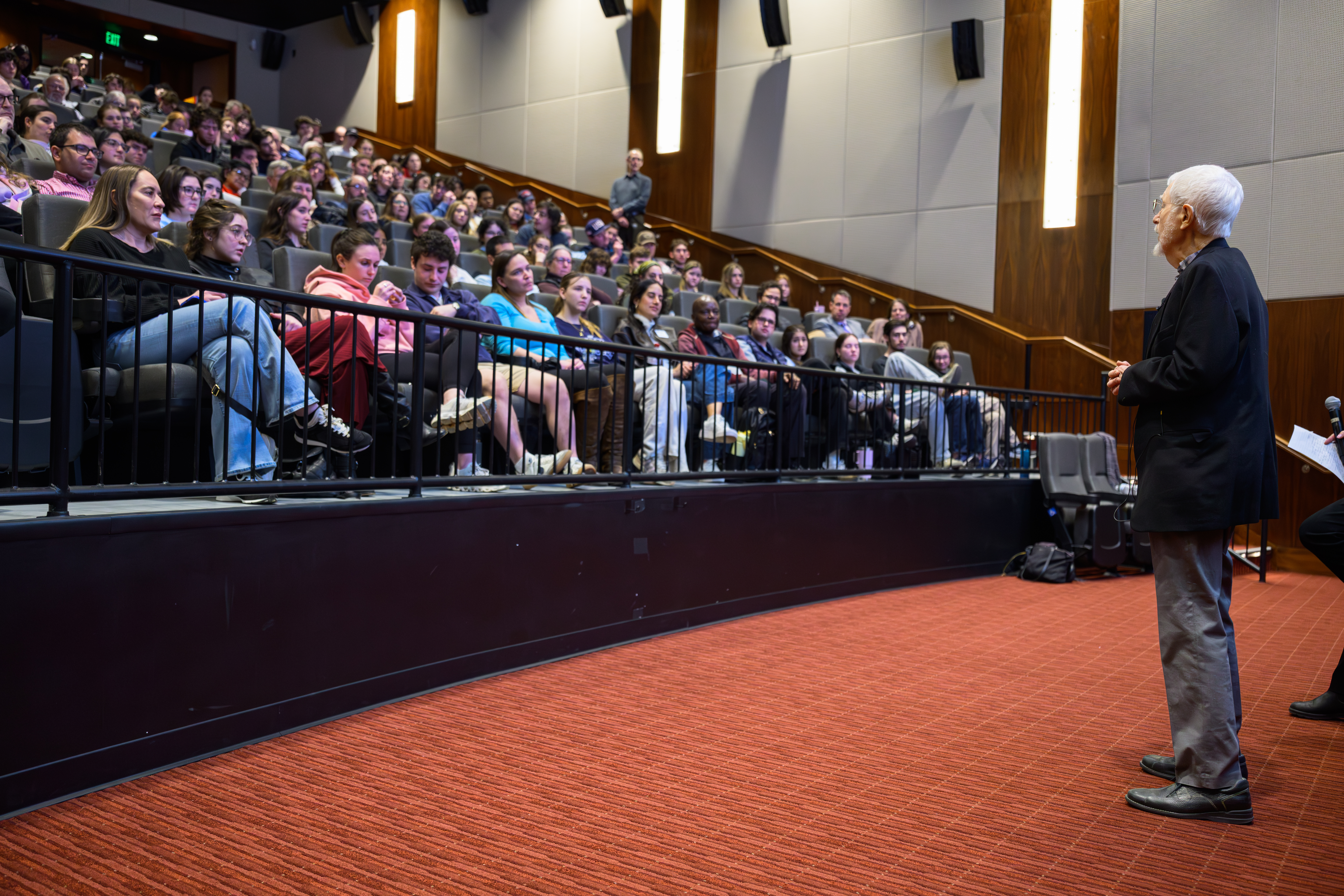 an older speaker stands at the front of the room facing rows of seated students during a formal presentation or remembrance event.