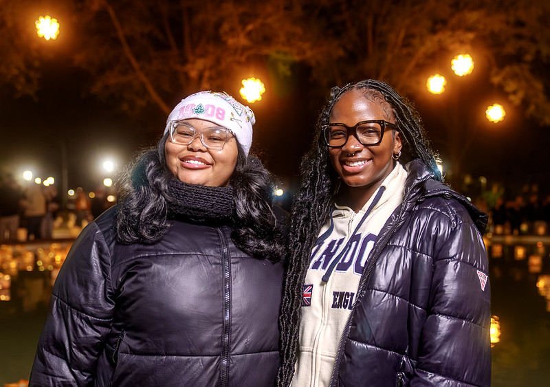 Two women wearing glasses and winter jackets smile at the camera while standing outdoors at night. Warm string lights glow in the background, illuminating trees and what appears to be a candlelit display behind them.