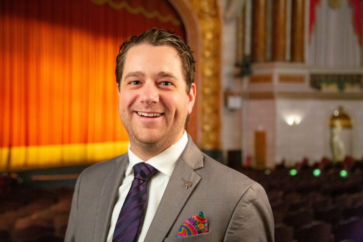 Scott Leighty in a suit and tie, smiling warmly at the camera inside a large theater.