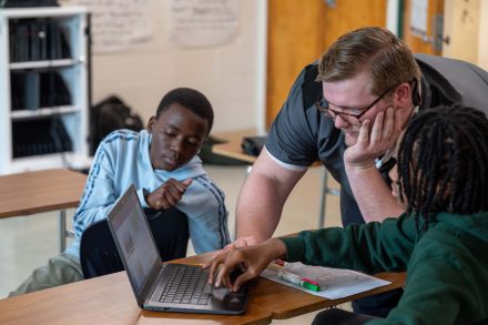 Two students sitting by a computer learning about AI from their teacher.