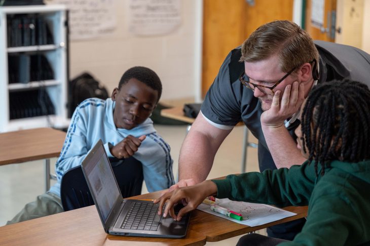 Two students sitting by a computer learning about AI from their teacher.