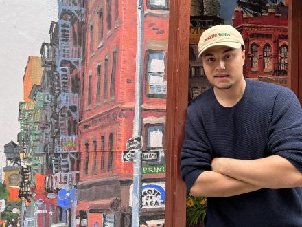 A man stands with arms crossed beside a colorful mural depicting an urban street scene with brick buildings. He wears a casual sweater and baseball cap, posing confidently outdoors.