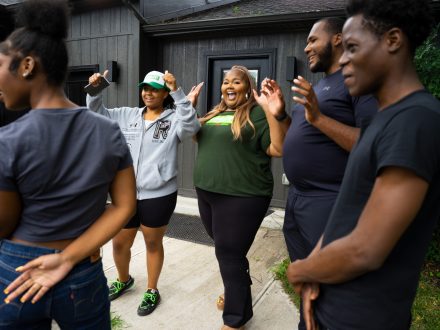 A group of people stand outdoors in front of a building, smiling, clapping, and raising their hands in celebration. The scene captures a joyful, supportive moment of shared excitement and community.