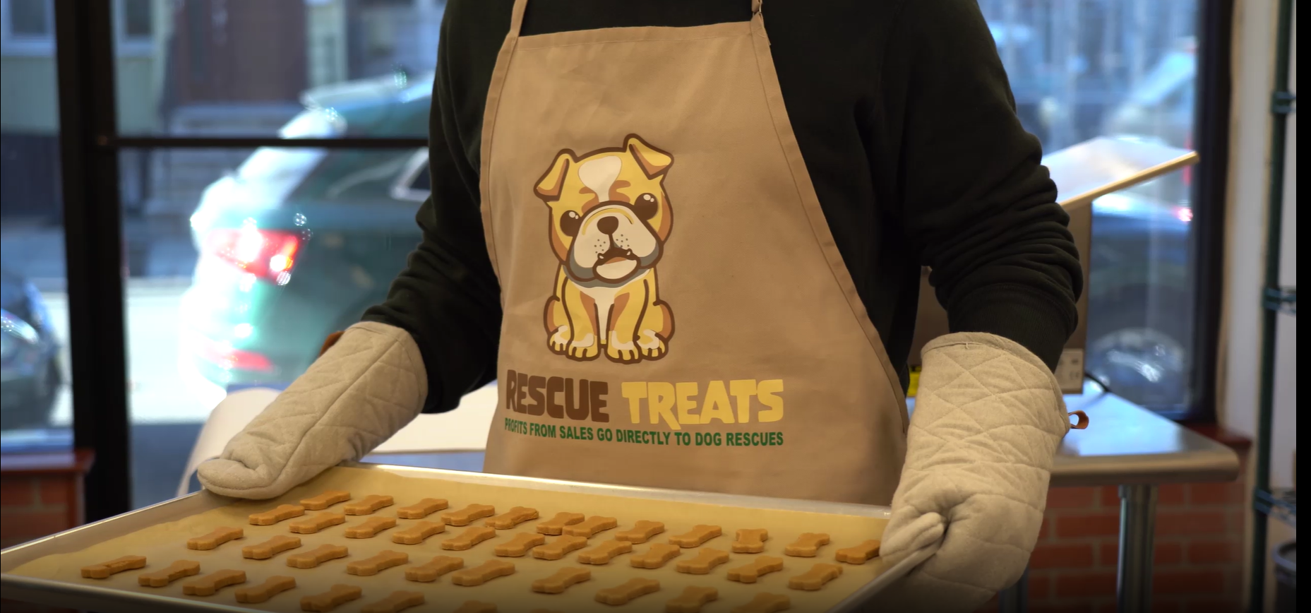 A person wearing oven mitts and a “Rescue Treats” apron holds a tray of bone-shaped dog treats inside a kitchen or bakery space. The treats are neatly arranged, highlighting a dog-focused baking activity.