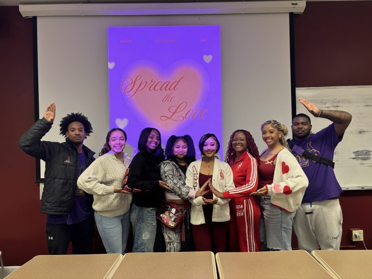 Eight students pose in a classroom beneath a “Spread the Love” Valentine’s display, forming a heart shape with their hands.