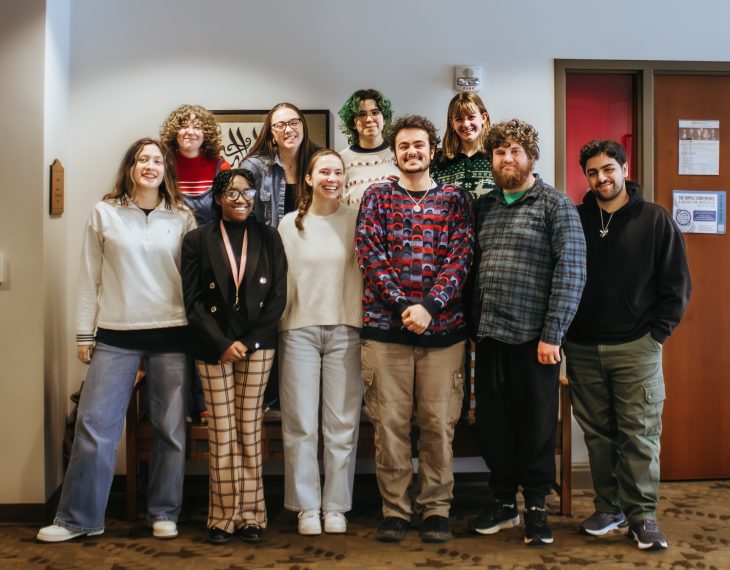 A group of students pose for a photo in front a white wall with a door to the right.