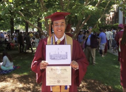 A graduate wearing a cap and gown smiles while holding a diploma and certificate outdoors during a commencement celebration. Other graduates and guests gather in the background under leafy trees.