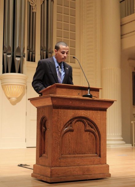 A student stands at a wooden podium in a formal auditorium, reading from prepared remarks. Large organ pipes and classical architectural details frame the stage.