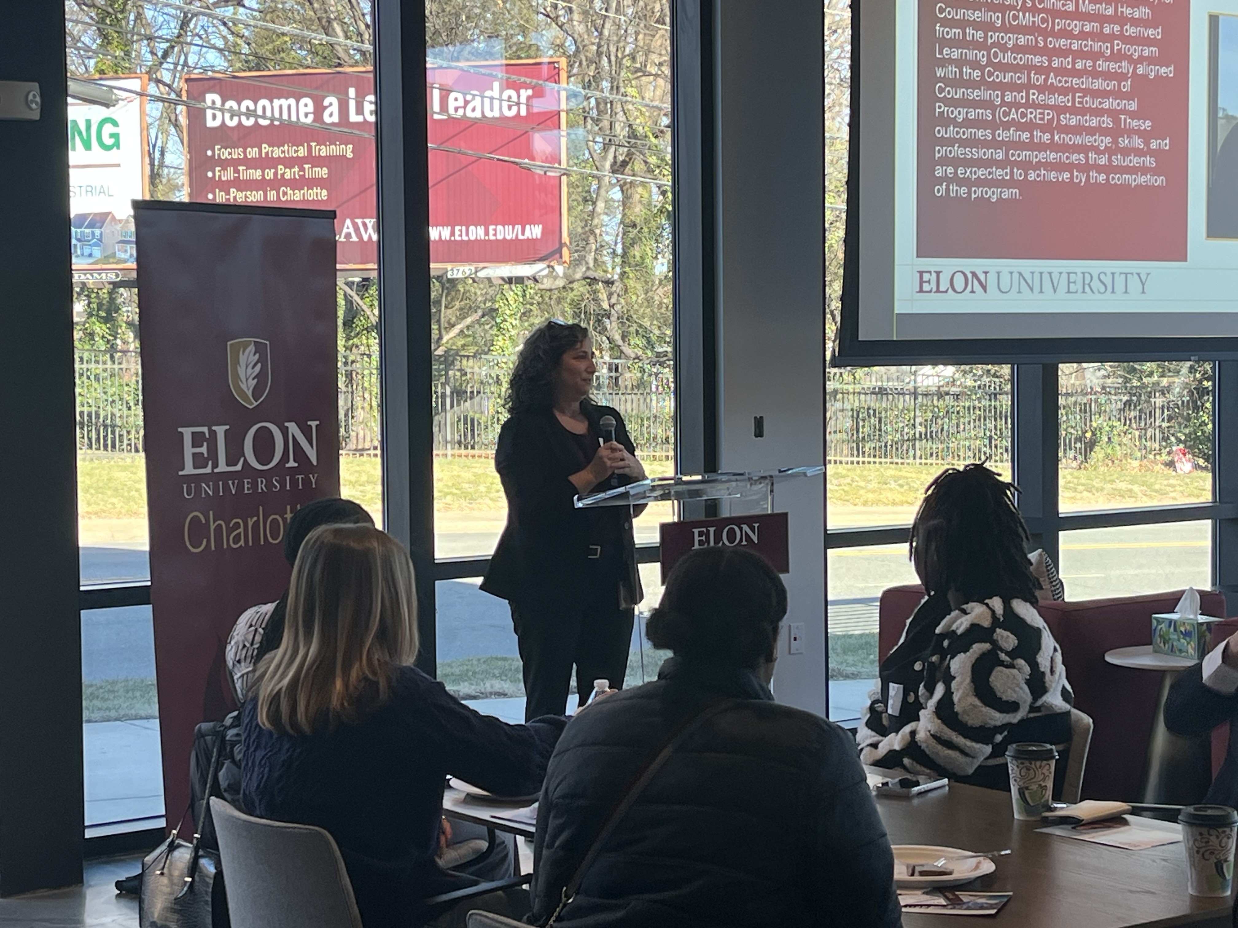 A speaker presents at a podium to a small audience during an Elon University Charlotte event, with a presentation slide displayed behind her.