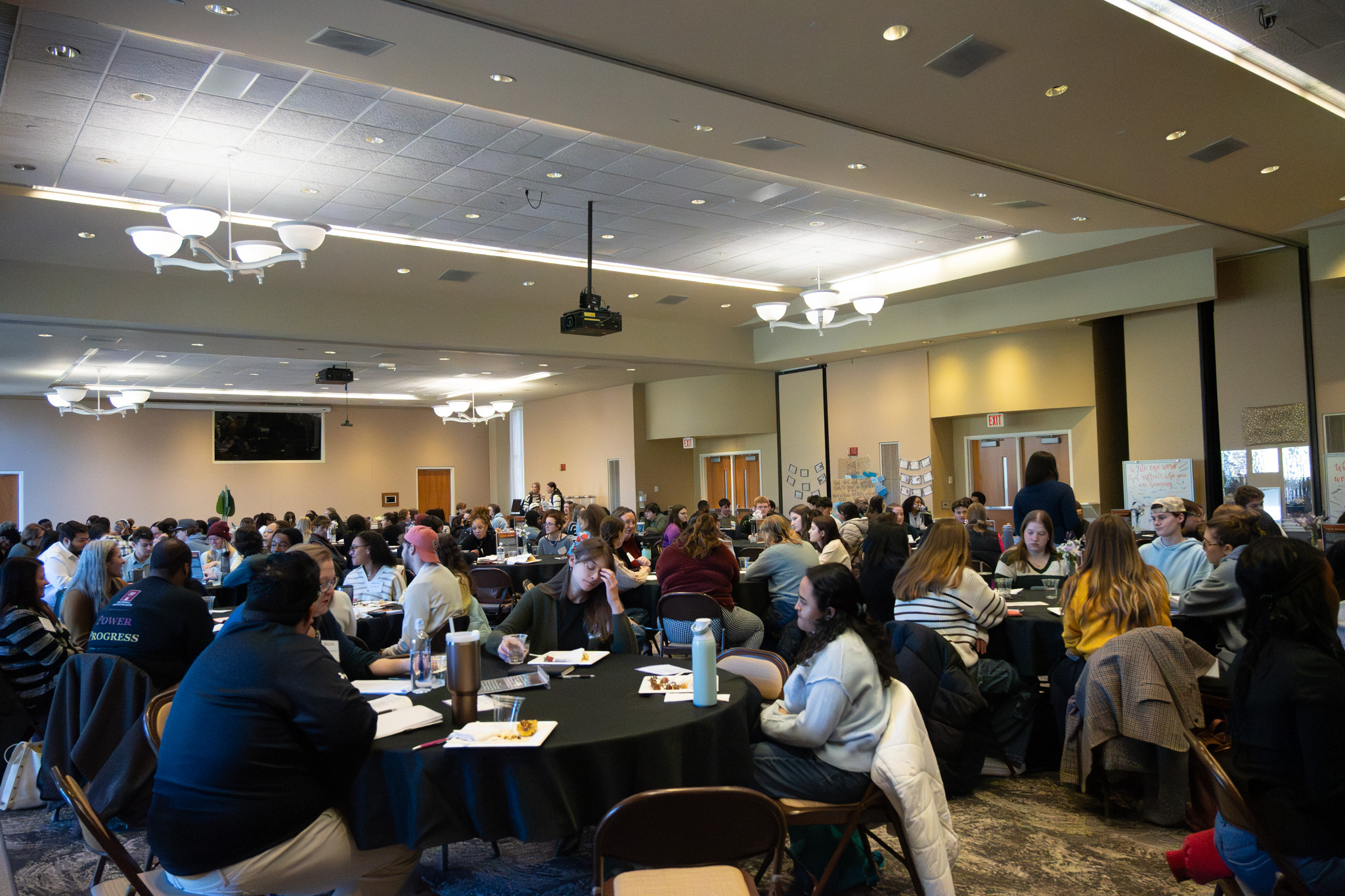 Students sit at round tables in a large conference room, engaged in discussion during a campus event.