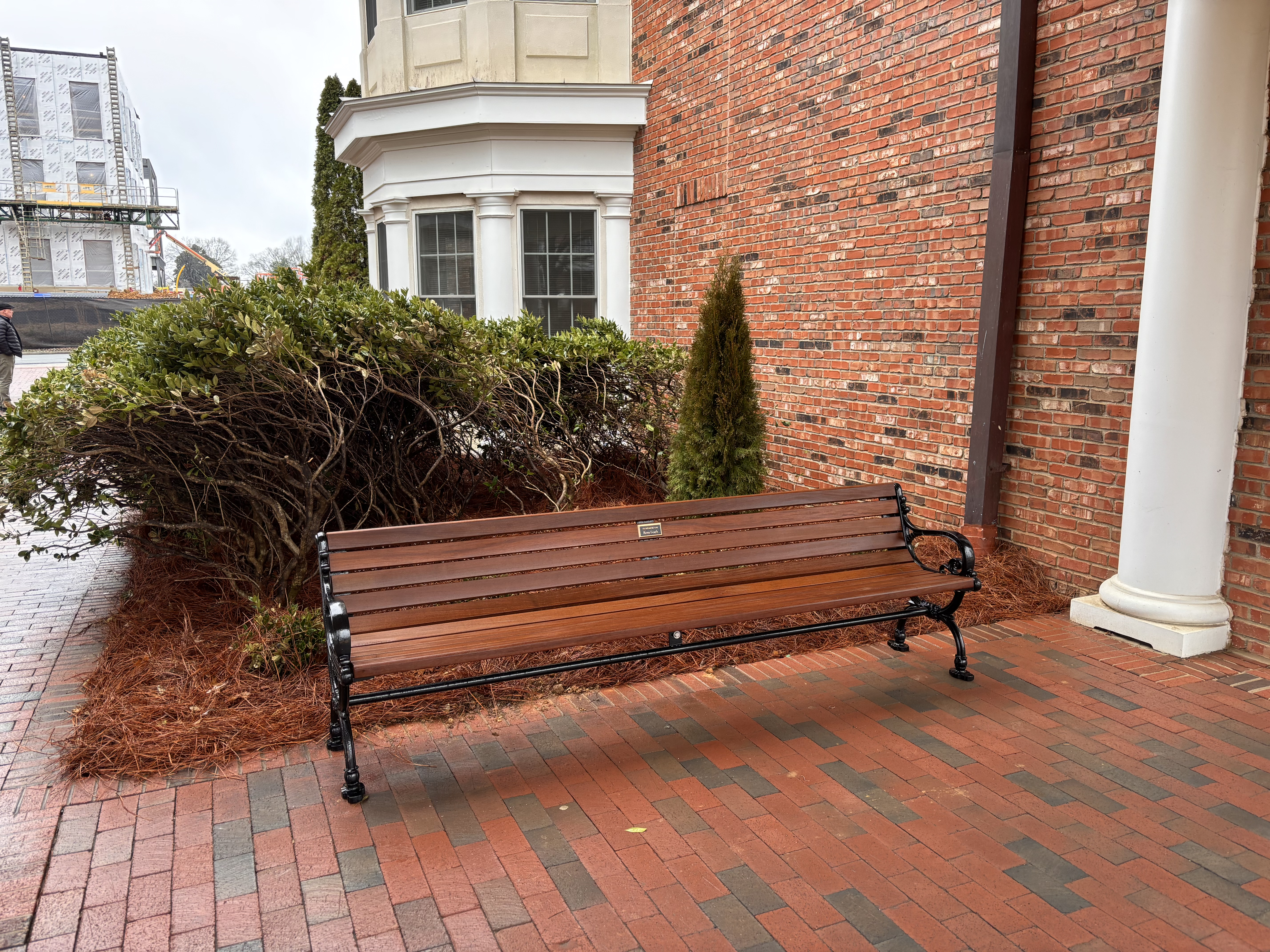 A bench sits next to a white column. There is construction in the background and a green bush.