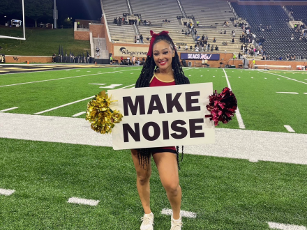 Maleah Proctor stands on a football field at night holding a “MAKE NOISE” sign with pom-poms in front of stadium stands.