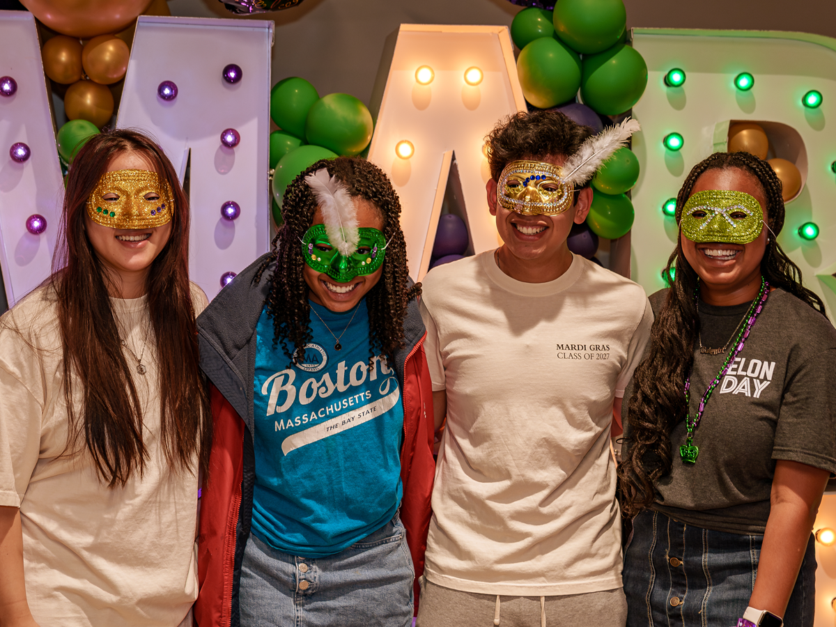 Four students wearing colorful Mardi Gras masks smile in front of illuminated “MARDI” letters and green and purple balloons.