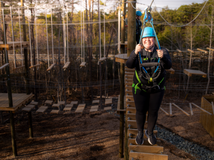 Student in helmet and harness smiles while balancing on a high ropes course element among trees.