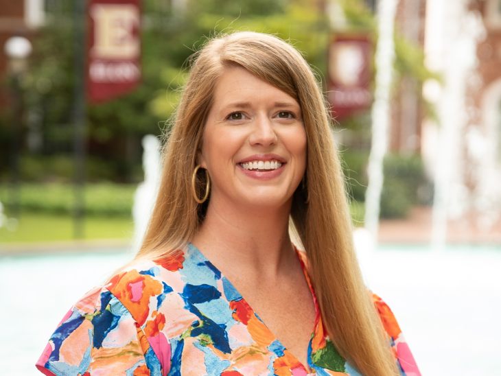 Sara Beth Hardy smiles in front of a campus fountain, wearing a colorful patterned blouse, with brick buildings and Elon banners in the background.