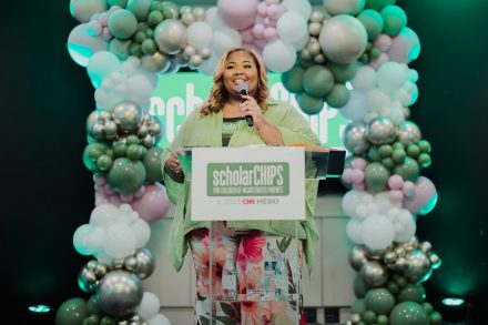 A person speaks at a clear podium labeled “scholarCHIPS – CHIP HERO” on a stage decorated with green, white, and metallic balloon arches, holding a microphone and smiling. The scene appears to be a formal recognition or awards event with professional lighting.