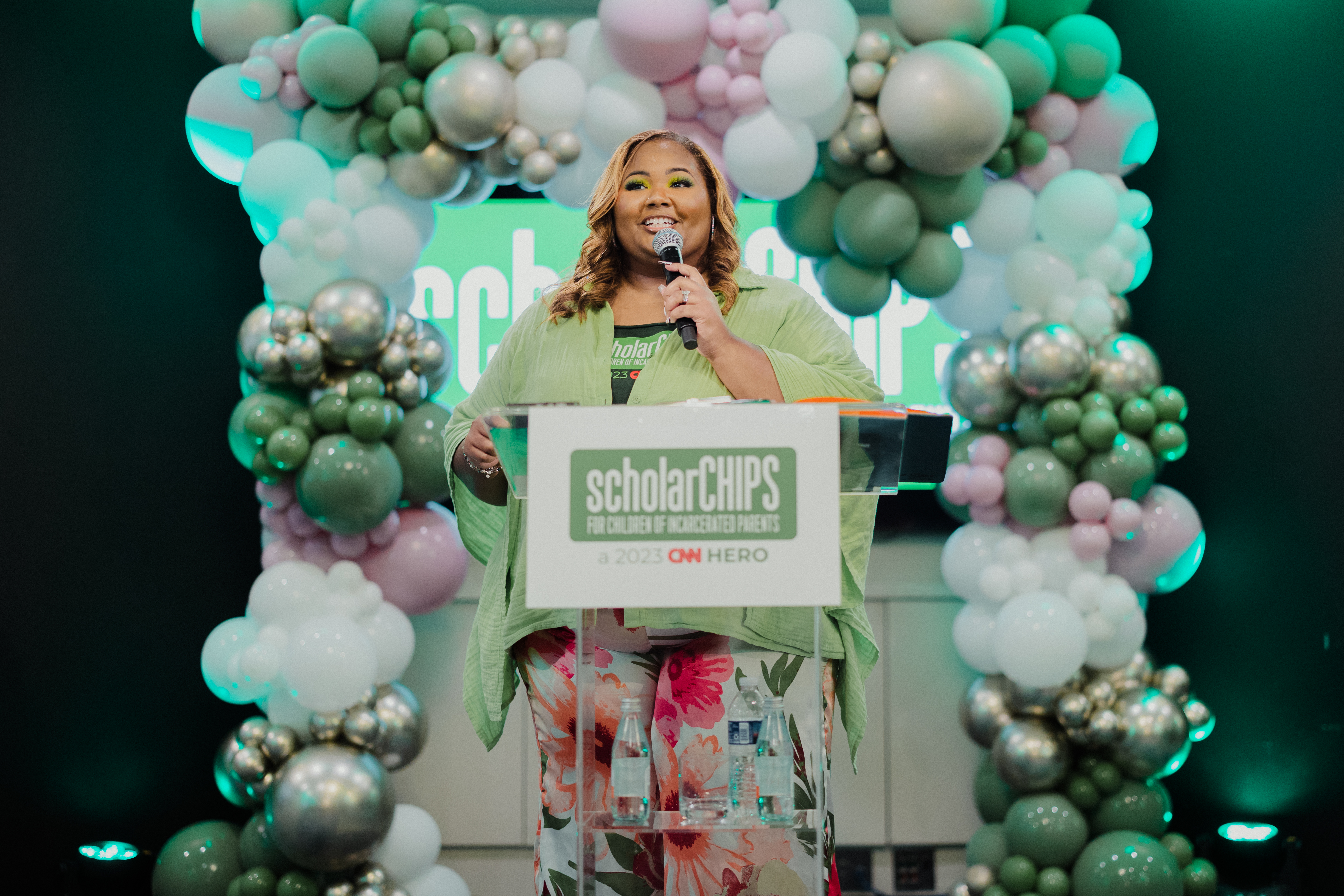 A person speaks at a clear podium labeled “scholarCHIPS – CHIP HERO” on a stage decorated with green, white, and metallic balloon arches, holding a microphone and smiling. The scene appears to be a formal recognition or awards event with professional lighting.