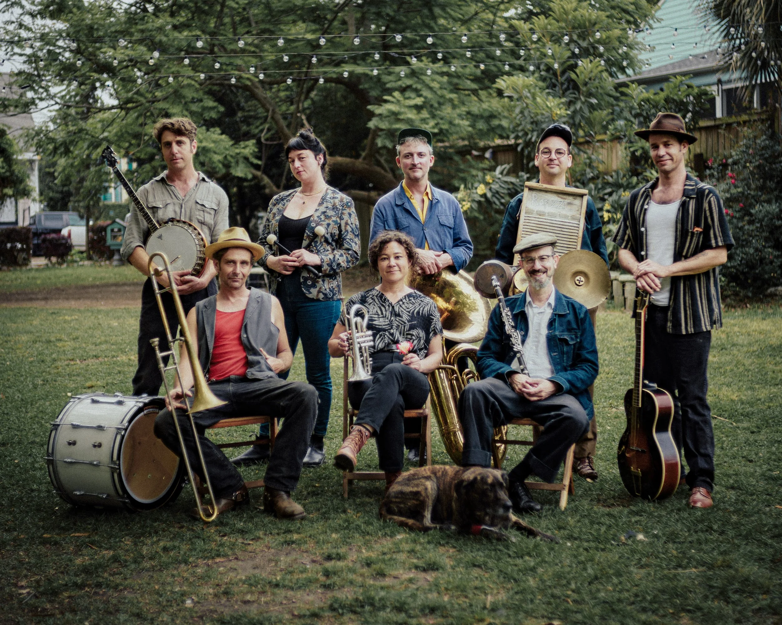 A group of musicians pose outdoors in a grassy yard, holding instruments including a banjo, trombone, trumpet, tuba, washboard, cymbals, and guitar. Some sit on chairs while others stand behind them, with string lights overhead and a dog lying in front of the group.