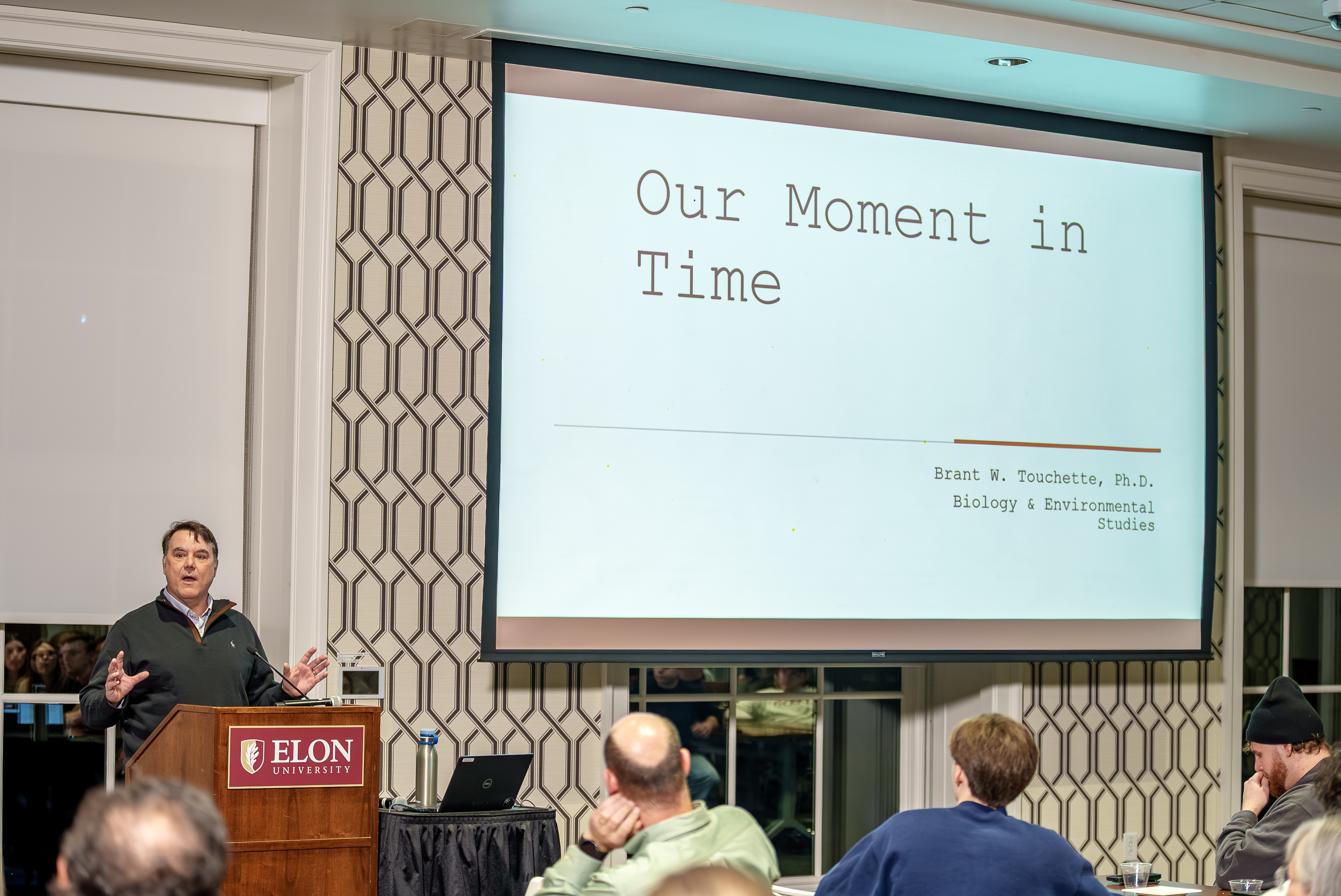A speaker stands at a podium with the Elon University logo, presenting to an audience while a large screen behind him displays a slide titled “Our Moment in Time.” Several attendees sit facing the stage, listening as he gestures with his hands during the lecture.
