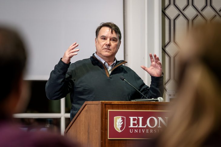 A man speaks at a podium with the Elon University logo, gesturing with both hands raised as he addresses an audience. Blurred figures in the foreground frame the scene, suggesting a lecture or public talk setting.