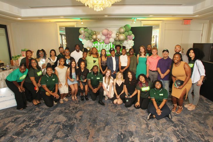 A large group of people pose together indoors in front of a “scholarCHIPS” backdrop with green, white, and metallic balloon decorations. The group appears dressed for a formal or celebratory event, smiling and facing the camera.