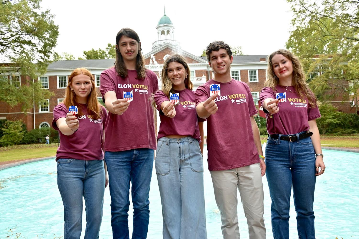 Five people maroon t-shirts stand in front of Fonville Fountain at Elon University. The shirt say 