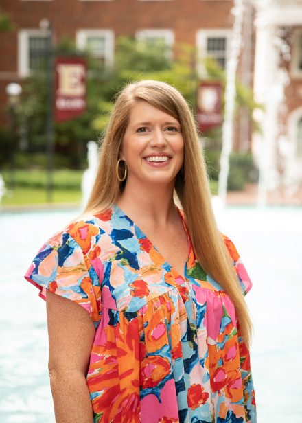 Sara Beth Hardy smiles in front of a campus fountain, wearing a colorful patterned blouse, with brick buildings and Elon banners in the background.